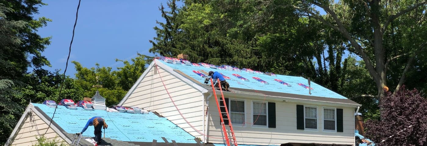 Roofing crew on a house mid-replacement, underlayment down and shingle bundles staged along the ridge
