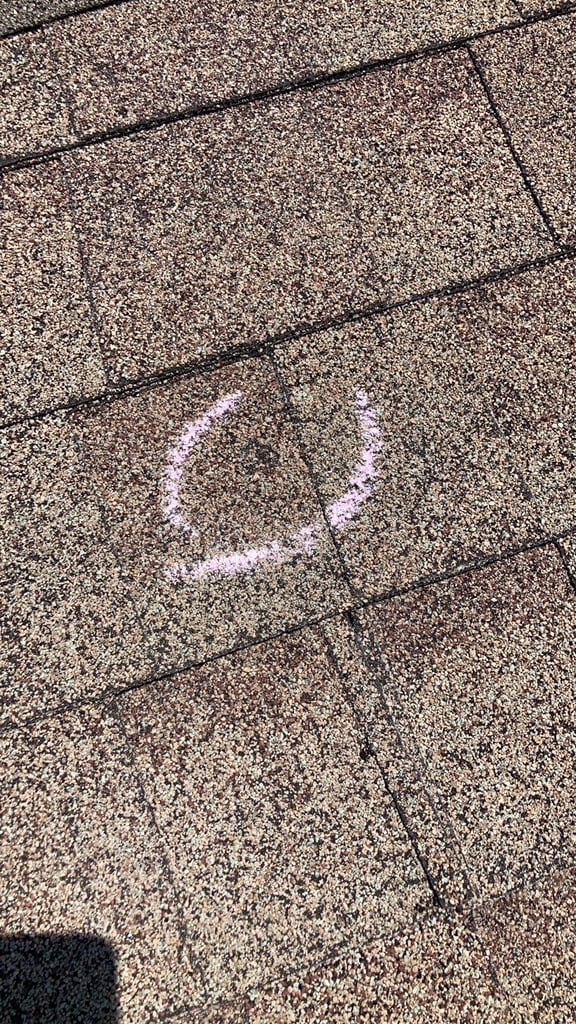 Single hail impact on an asphalt shingle circled in chalk, granules crushed away at the centre